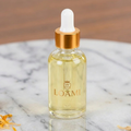 A clear glass dropper bottle filled with pale yellow liquid, featuring a metallic gold collar and a white rubber dropper top. The bottle is printed with the brand name 'LOAMI' and is set on a white marble surface with dried orange petals.